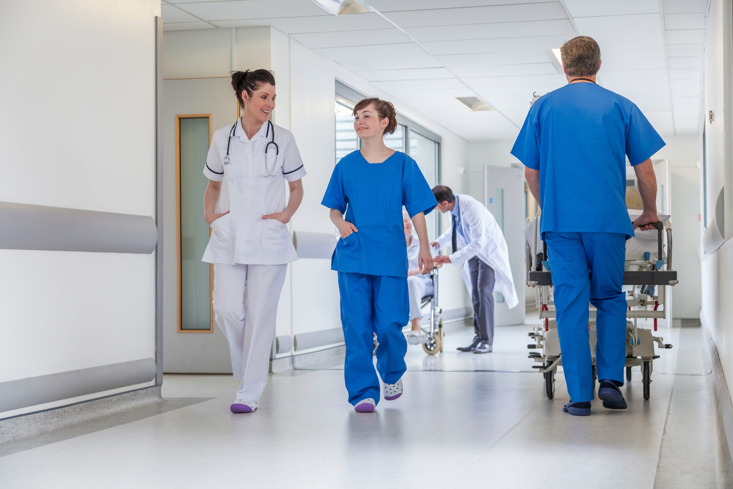 Male nurse pushing stretcher gurney bed in hospital corridor with male & female doctors & nurses & senior female patient in a wheelchair